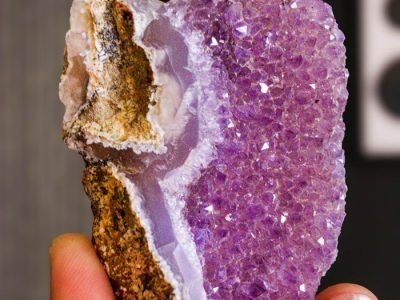 Home Detailed view of a purple amethyst geode crystal held between fingers, showcasing natural beauty.